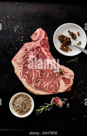 Raw cowboy Rindersteak mit Gewürzen Zutaten, Spice Rub und Kräutern auf einem schwarzen Hintergrund gesehen als Flach Stockfoto