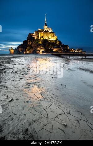 Nachtaufnahme der französischen Festung Le Mont-Saint-Michel, deren Lichter sich auf dem grauen Ton im Vordergrund spiegeln, gegen einen blauen Himmel Stockfoto