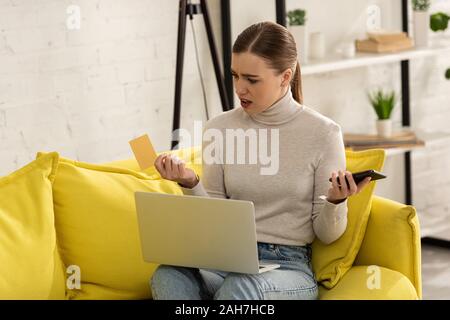 Verwirrt Frau mit Kreditkarte, Smartphone und Laptop auf dem Sofa zu Hause Stockfoto