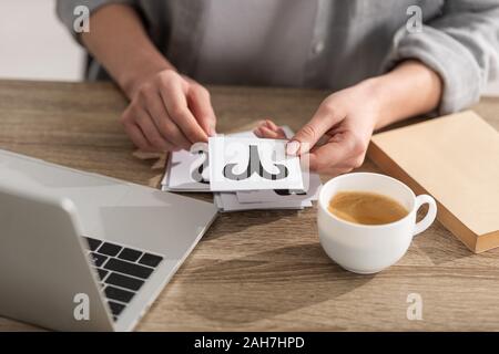 7/8-Ansicht von Astrologen halten Karten mit Sternzeichen neben Laptop und Kaffee auf Tisch Stockfoto