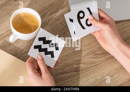 7/8-Ansicht von Astrologen halten Karten mit Sternzeichen neben Kaffee und Buch auf Tabelle Stockfoto