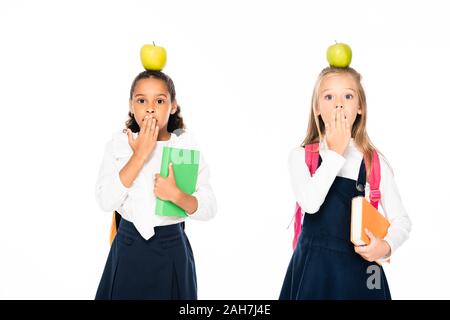 Zwei schockiert multikulturelle Schülerinnen mit Äpfeln auf Kopf, Mund mit den Händen isoliert auf weißem Stockfoto