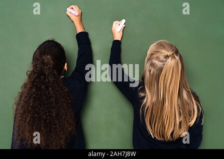 Zurück Blick auf zwei multikulturelle Schülerinnen schreiben auf der Schiefertafel mit Kreiden Stockfoto