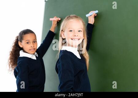 Zwei lächelnde multikulturelle Schülerinnen schreiben auf der Schiefertafel, während bei Camera suchen Stockfoto
