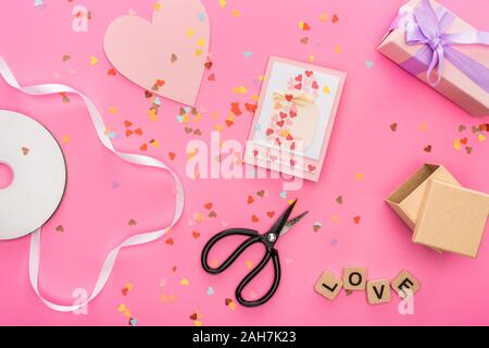 Blick von oben auf die valentines Konfetti, leere Compact Disk, Schere, Geschenkboxen, Grußkarte und liebe Schriftzüge auf holzwürfel auf rosa Hintergrund Stockfoto