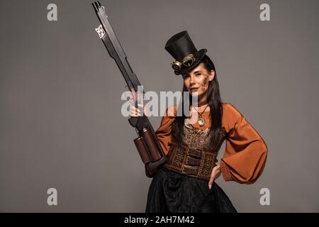 Attraktive steampunk Frau in Hut holding Gun und stehen mit Hand auf die Hüfte auf Grau isoliert Stockfoto