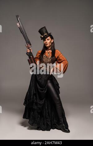 In voller Länge ansehen attraktiver steampunk Frau in Hut holding Gun und stehen mit Hand auf die Hüfte auf Grau Stockfoto