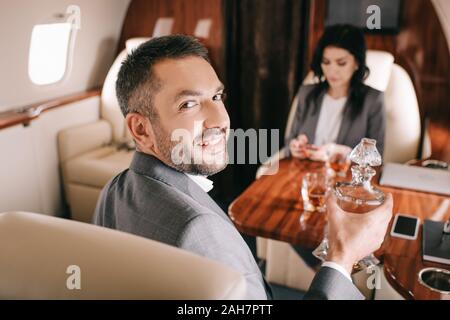 Selektiver Fokus der glücklichen Geschäftsmann holding Glas mit Alkohol in der Nähe von geschäftsfrau in Private Jet Stockfoto