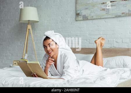 Attraktive Frau in ein Handtuch und Bademantel holding Buch am Morgen Stockfoto