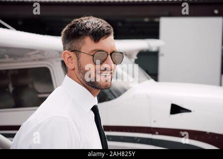 Bärtige Pilot in formale Abnutzung und Sonnenbrille lächelnd in der Nähe der Ebene Stockfoto