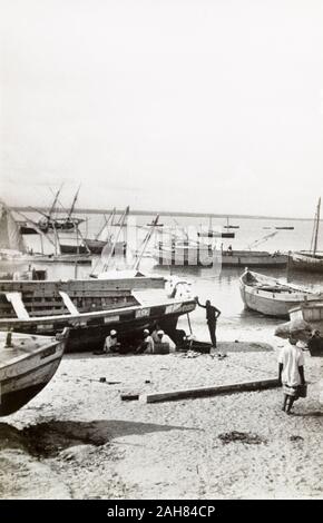 Kenia, einer Gruppe von Männer sitzen am Strand neben einem trocken-Boot im Hafen von Lamu angedockt. Es gibt mehrere andere Fischerboote im Wasser und am Strand. Die ursprünglichen Manuskript Bildunterschrift: Lamu, 1947. 1995/076/1/1/13/8. Stockfoto