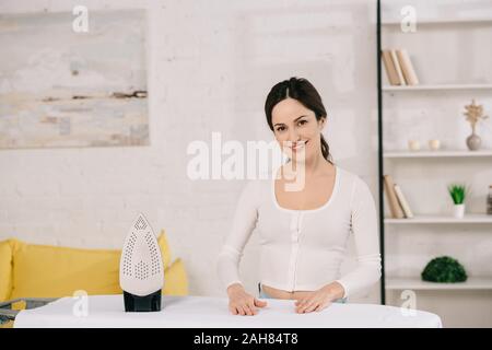 Hausfrau glücklich lächelnd in die Kamera beim Stehen in der Nähe von Bügelbrett Stockfoto