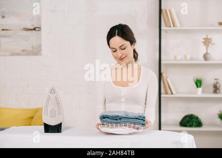 Junge Hausfrau Holding gebügelt und lächelnd beim Stehen in der Nähe von Bügelbrett Stockfoto