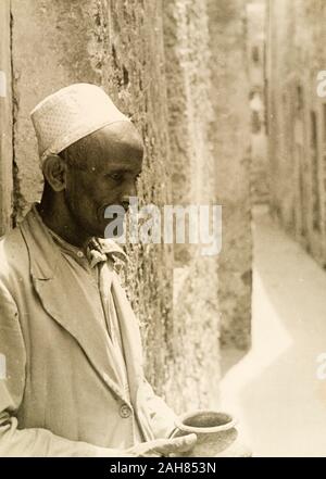 Kenia, Profil, geschossen von einem arabischen Mann, eine gestickte kufi (Gap). Er lehnt sich gegen eine Mauer aus Stein auf einer schmalen Straße, einen Tontopf. Das Foto ist ein Duplikat eines anderen in der Sammlung, die das ursprüngliche Manuskript Titel' einen arabischen Schmied, der an der Tür der sein Geschäft in einer der engen Gassen von Lamu, an der Küste von Kenia.', 1947. 1995/076/1/2/7/14. Stockfoto