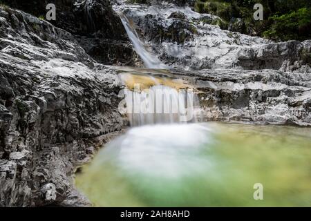 Niedriger Winkel Blick auf einen Wasserfall in den Bergen, umgeben von grüner Vegetation, mit einem Strom von Wasser, der über weißem Fels und in einen grünen Teich fließt Stockfoto