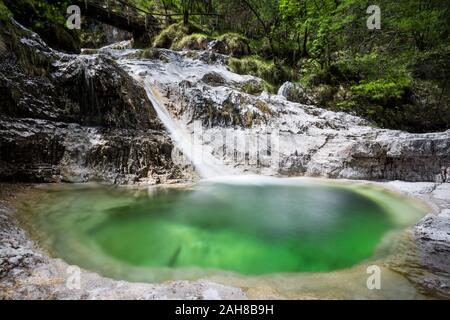 Weitwinkelansicht eines alpinen Wasserfalls, umgeben von Vegetation, mit einem Wasserfluss, der zwischen weißen Felsen und in einen smaragdgrünen Teich fließt Stockfoto