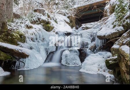 Weitwinkelansicht der Winterlandschaft, mit einem kleinen Wasserfall, der zwischen eisbedeckten Felsen und in einen kristallklaren Teich fließt, umgeben von Pinien Stockfoto