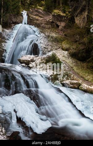 Weitwinkelansicht eines gefrorenen Wasserfalls in norditalien, umgeben von eisbedeckten Felsen und Pinien Stockfoto
