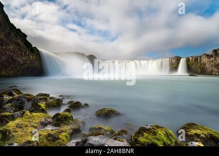 Weitwinkelansicht des isländischen Wasserfalls von Godafoss, mit moosbedeckten Felsen im Vordergrund, unter einem blauen Himmel mit geschwollenen Wolken Stockfoto