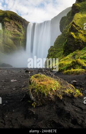 Niedriger Blickwinkel auf den riesigen Wasserfall von Skogafoss, der zwischen moosbedecktem Felsen und einem schwarzen Sandstrand fließt Stockfoto