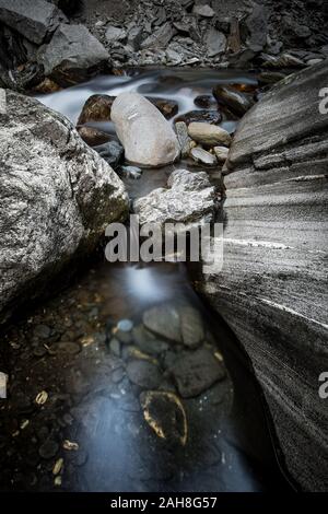 Weitwinkelblick auf einen Bergbach, der zwischen Felsen und Kieselsteinen fließt und in einen kristallklaren Teich mündet Stockfoto