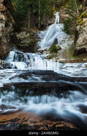 Niedriger Blick auf einen tiefgefrorenen, alpinen Wasserfall zwischen Felsen und aus Kiefernholz Stockfoto