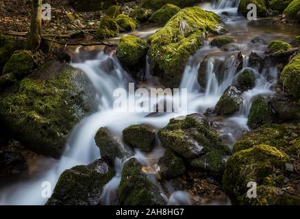 Weitwinkelansicht einer Berglandschaft mit einem Bach, der durch eine Reihe von Stromschnellen zwischen moosbedeckten Felsen fließt Stockfoto