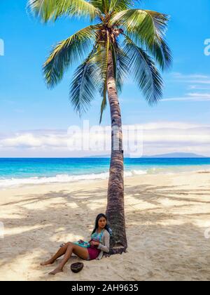 Insel Mindoro, Philippinen - Februar 16., 2011. Eine junge asiatische Frau sitzt unter einer Palme am Strand. Stockfoto