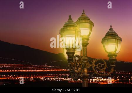 Ein Vintage street lamp glühen hell mit die beleuchtete Stadt und die Berge im Hintergrund nach dem Sonnenuntergang. Stromverbrauch Konzept. Stockfoto