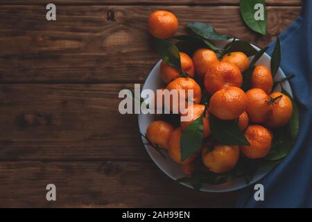 Mandarinen mit Blättern in einer Platte auf einem hölzernen Hintergrund. Ansicht von oben, Platz für Text. Stockfoto