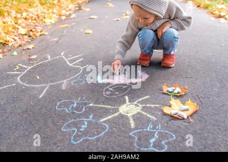 Kleiner Junge von 3-5 Jahren, sitzt auf dem Bürgersteig und zeichnet Zeichnungen mit bunten Buntstiften, Freiraum für Kreativität, Kinder. In warmen Kleidern im Herbst Stockfoto