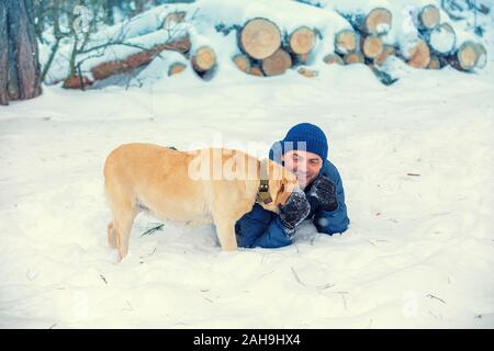 Ein Hund und Mensch sind die besten Freunde. Eine glücklich lächelnde Mann liegen auf dem Schnee im Winter. Der Mann spielt mit einem Labrador Retriever Hund Stockfoto