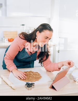 Junge brünette Frau, die ein Weihnachtsplätzchen zu Hause mit einem Tablet das Rezept zu prüfen. Handgemachte Lebkuchen Cookies. Stockfoto