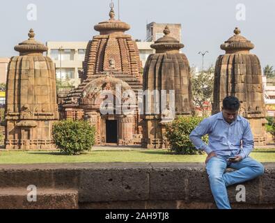 Bhubaneshwar, Orissa, Indien - Februar 2018: Ein Mann sitzt an einer Steinmauer und starrt mit den alten Mukteshwar Tempeln im Hintergrund an seinem Telefon. Stockfoto