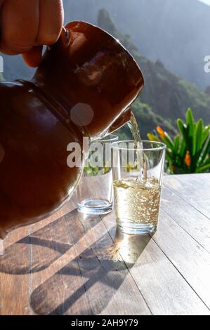 Man gießt Weißwein aus Ton Kanne in Glas auf der Terrasse in sunlights mit Blick auf die grüne Landschaft der kleinen Bergdorf Masca auf Teneriffa, Spa Stockfoto