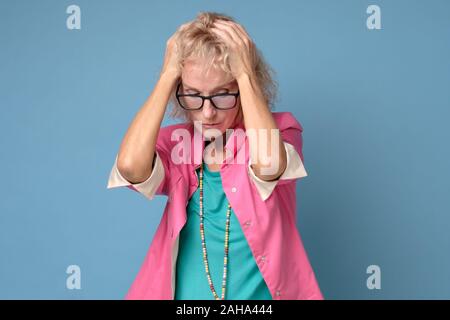 Europäische reife blonde Frau in Gläsern zum Ausdruck der Frustration in Panik. Studio Schuß auf blauen Wand Stockfoto