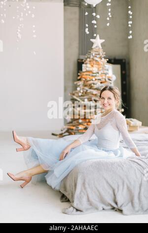 Hübsche junge Frau in Blau-weiß Kleid sitzt auf dem Bett, auf dem Hintergrund der Weihnachtsbaum. Stockfoto