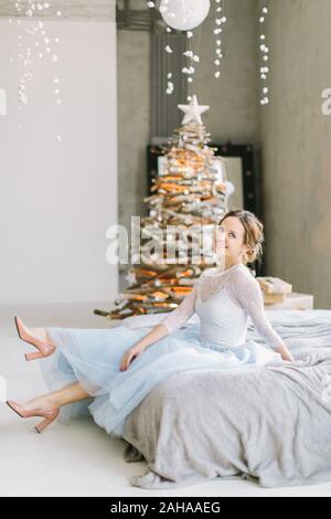 Hübsche junge Frau in Blau-weiß Kleid sitzt auf dem Bett, auf dem Hintergrund der Weihnachtsbaum. Stockfoto