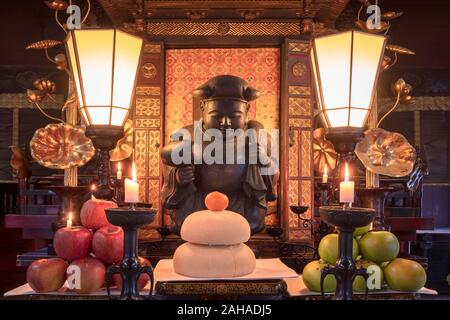 Tokyo, Japan - 7. Januar 2020: mochi's neue Jahr Reis, Clementine und Äpfel Angebote vor eine hölzerne Statue der Daikokuten japanische Gottheit. Stockfoto