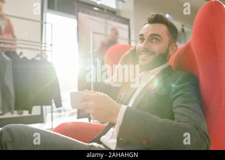 Junge erfolgreiche Geschäftsmann mit Tasse Tee consulting Client per Handy Stockfoto
