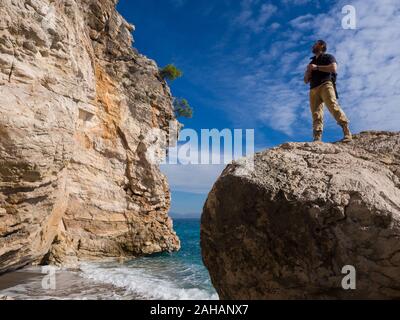 Reisende mit Rucksack auf den Felsen in der Nähe von dem Meer entfernt. Sommer Reisen Urlaub. Hübscher junger kaukasischen touristische Mann in legere Kleidung im Freien Stockfoto