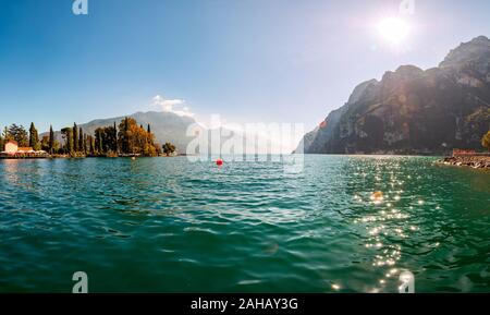 Panoramablick gegen die hellen Sonnenstrahlen oberhalb der Felsen am wunderschönen Gardasee Ufer in Riva del Garda Stadt in der Lombardei, Italien, umgeben von hohen Dol Stockfoto