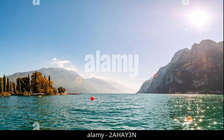 Panoramablick gegen die hellen Sonnenstrahlen oberhalb der Felsen am wunderschönen Gardasee Ufer in Riva del Garda Stadt in der Lombardei, Italien, umgeben von hohen Dol Stockfoto