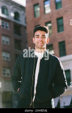 Smiling hispanic Männlich schaut direkt in die Kamera, während in der Stadt an einem sonnigen Tag - Zufrieden mit dem Gebäude im Hintergrund Stockfoto