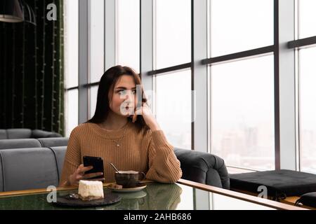 Ein Mädchen sitzt in einem Café, hält ein Handy in der Hand und schaut aus dem Fenster und dachte an etwas Stockfoto