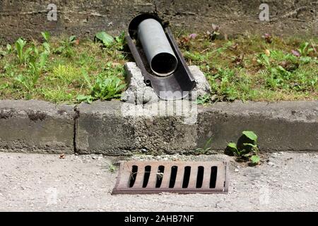 Behelfsmäßige Kunststoff und Metall Dachrinne mit Beton und Gras führende Wasser aus Steinmauer umgeben zu Storm drain Rinne auf asphaltierten Straße Stockfoto