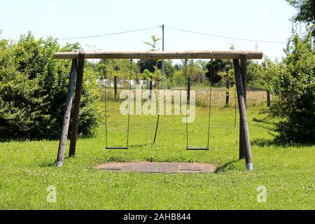 Alte verfallene hölzerne Retro Vintage outdoor öffentlichen Spielplatz ausrüstung in Form von Swing mit zwei Sitze mit ungeschnittenem Gras und niedrigen Wald umgeben Stockfoto