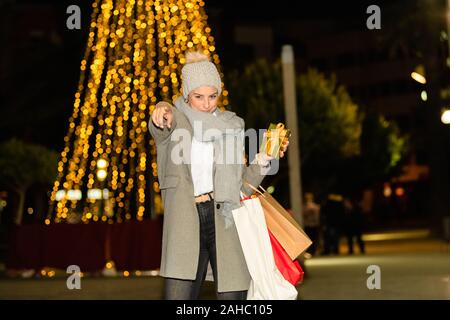 Lächelnde Frau mit Papier Tüten und halten eine goldene verpackt Geschenkkarton Punkte an der Kamera auf eine unscharf Hintergrund. Einkaufsmöglichkeiten für Geschenke co Stockfoto