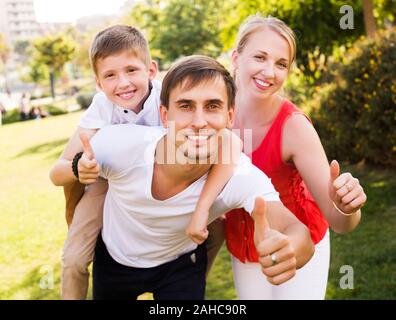 Glückliche junge Eltern mit Jungen in der Schule Alter Spaß und zeigen Daumen hoch im Freien Stockfoto