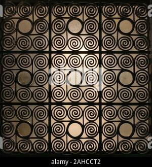 Griechische Wrought Iron Gate Gitter, verziert mit kunstvoll verdrillte Spiralen und mit Milchglas im Hintergrund, dass leicht beleuchtet wird. Stockfoto